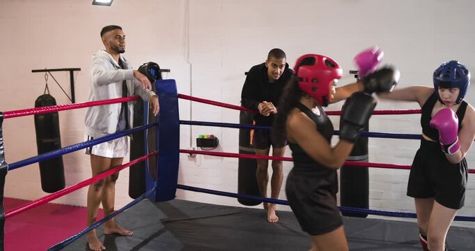 Coach raising arm signaling female fighters wearing headgear sparring in ring jab kick combos