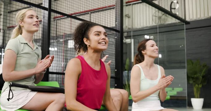 Diverse female friends reacting to play on green bleachers by leaning forward, clapping, smiling