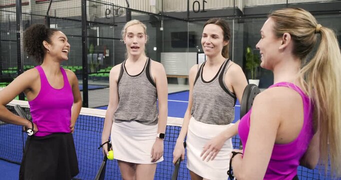 Diverse female friends holding rackets chatting smiling on padel court huddling by net for unity