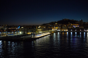 Obraz premium Panorama of Naples, Italy, at night. The city lights of a coastal port glitter against the dark night sky, reflecting beautifully on the calm sea surface. There are buildings and a distant hill.
