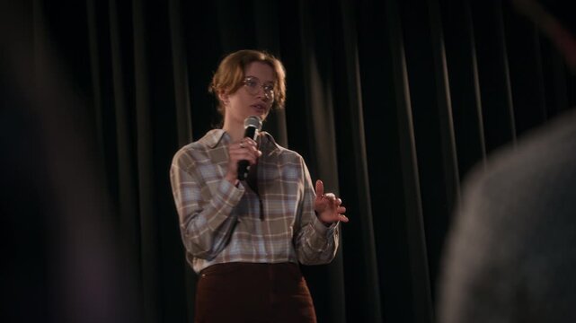 Shot from the audiences point of view of young adult female comedian with glasses setting joke then landing punchline while performing on low key lit stage of comedy club, copy space