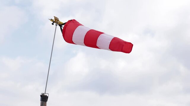 Low angle shot of a red and white striped windsock inflated by the wind, mounted on a pole with a small airplane ornament against a cloudy sky