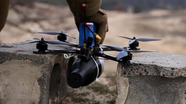 Horizontal close-up of a soldier's gloved hand preparing a tactical FPV kamikaze drone with a mounted explosive payload on a concrete block for mission