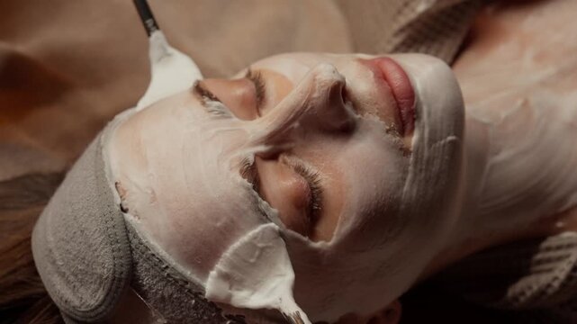 Relaxing facial treatment with creamy mask applied by beautician using soft brushes on peaceful woman lying with closed eyes in soothing spa wellness setting