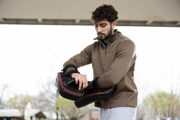 Young man wearing a quarter-zip jacket, standing outdoors, and putting on target mitts for a boxing...