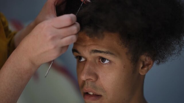 Close-up of a hairdresser's hands meticulously styling a young mixed-race man's afro hair, who shows expressions of discomfort and concern