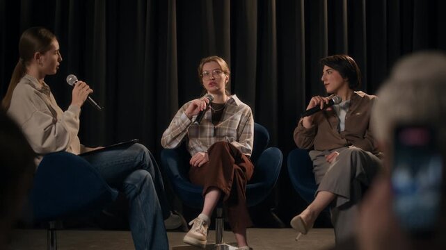 Rack focus shot of unrecognizable spectator recording video on smartphone of three young adult female comedians performing panel style stand up comedy on stage
