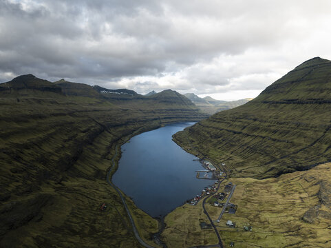 Aerial view of a deep blue fjord surrounded by steep green mountains and a small coastal village under a cloudy sky in Leynar, F