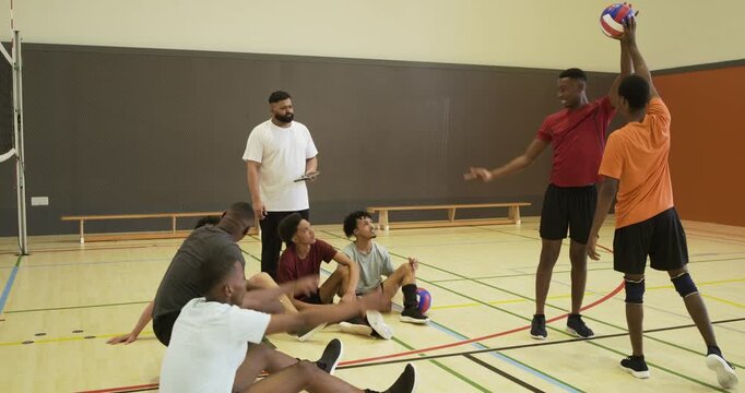 Diverse volleyball team and coach with tablet coaching at net, player holding ball, blocker leaping