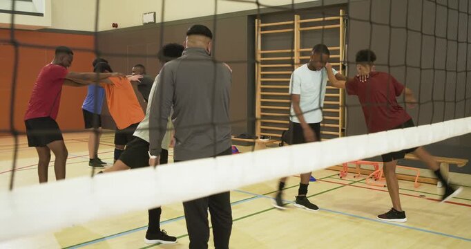 Male players warming up with leg swings by net in school gym after coach cueing now
