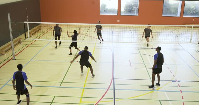 Two male volleyball teams in shirts in school gym, server serving ball, teams rallying to score