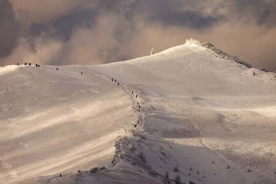 Close-up of hikers trekking on a snowy mountain ridge in Bieszczady during winter at sunset with dramatic clouds in the background.