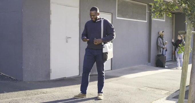 African American man walking sidewalk by gray wall holding cup messenger bag reading phone kicking