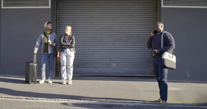 Diverse pair waiting by suitcase at shutter while man following phone call walking and gesturing