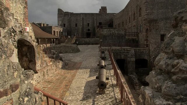 Interior courtyard of Citadelle Laferri&egrave;re fortress in Haiti, featuring rusty cannons lined up on cobblestone paths, stone walls, arches, and overcast sky above historic ruins.