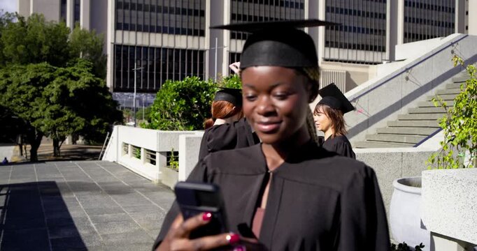 Female grad gowned reading phone message smiling holding diploma posing for grad portrait on campus