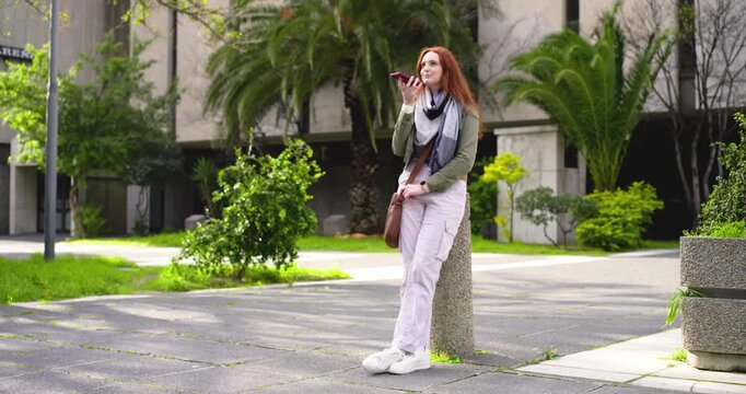 Redheaded woman leaning on bollard at campus plaza starting voice note on phone as passerby crosses