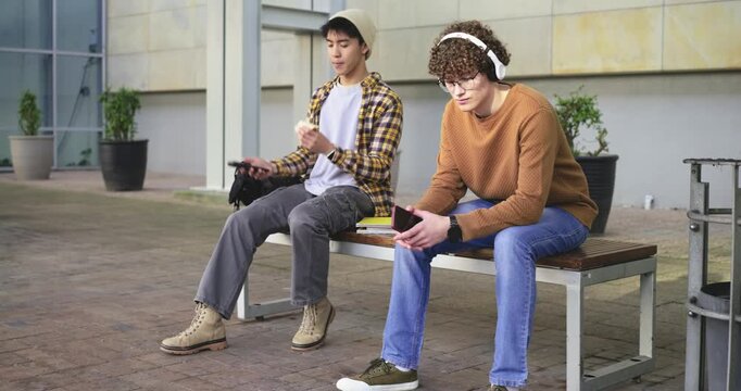 Diverse males sitting on campus seat talking on phone eating listening on headphones glancing left