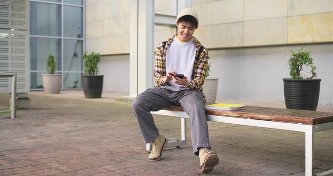 Asian man wearing hat and plaid sitting on bench in courtyard, checking messages, turning at walker