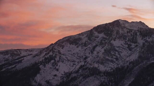 Fotage of a rugged snow-covered mountain during sunset. The peaks are highlighted by warm hues of orange, pink, and purple as the sun dips below the horizon. Dark patches of trees and rock