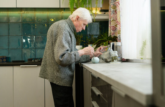 Side view of a senior citizen with hunched back preparing tea. The image illustrates challenges of aging and determination to maintain daily routines. Grandpa in kitchen.
