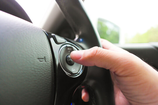 Car Cruise Control Buttons, A close-up of a driver's hand pressing the SET and RES cruise control buttons on a leather-wrapped steering wheel for a steady highway speed.