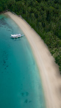 Aerial view of a traditional bangka boat anchored on a pristine white sand beach next to a lush palm tree forest and turquoise o