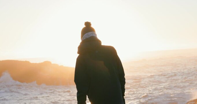Cinematic silhouette of person in knit beanie and winter coat standing by ocean during golden hour sunset. Warm amber light illuminates waves and mountains in background. Peaceful contemplative mood