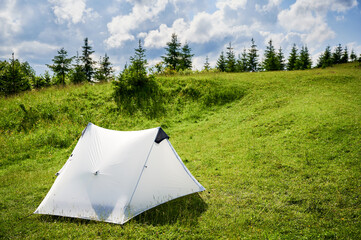 Naklejka premium White tourist tent on sunlit, grassy hilltop, surrounded by young pine trees. Panoramic view of lush, rolling mountains under bright blue sky with scattered clouds, creating idyllic camping spot.