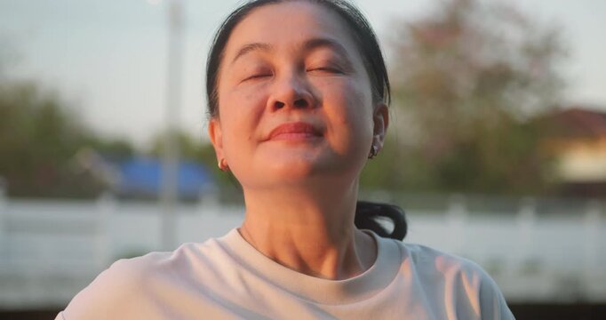 Middle aged Asian woman closing eyes and breathing fresh air in the park at sunset.