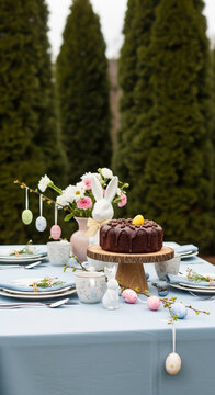 Outdoor Easter table with chocolate cake, floral arrangement, ceramic rabbit, eggs. Festive scene celebrating spring, renewal, Easter holiday season