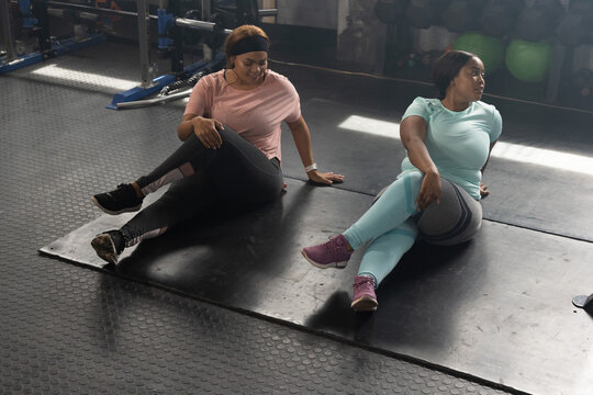 African American women performing seated spinal-twist stretches on black mats in gym weight area