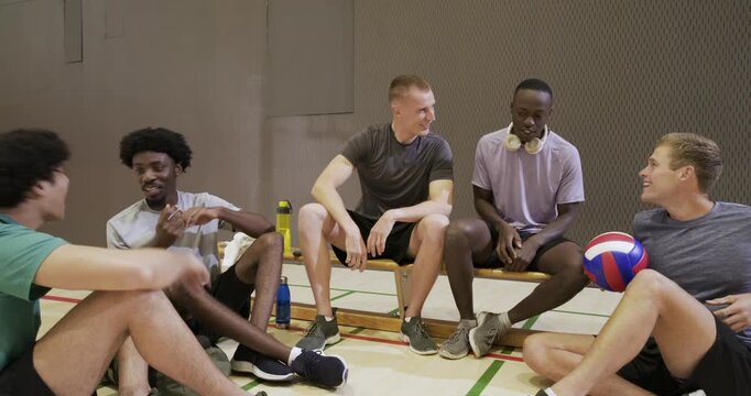 After practice diverse male athletes resting on bench at gym court, passing volleyball and laughing