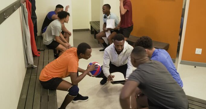Diverse male volleyball team in sportswear gathering, coach showing tablet, huddling to play