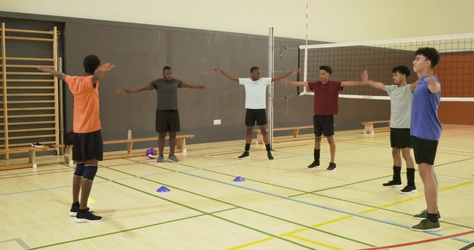 Diverse players following gesturing coach in orange shirt, raising arms warmup in gym by net, cones