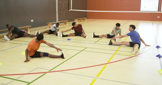 Diverse male volleyball team sitting on court reaching toes, following coach, warming up with ball