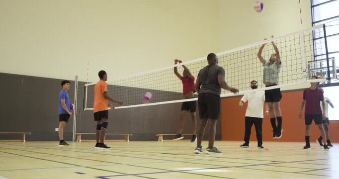 Male players, coach practicing in school gym, blue player initiating play over net with purple ball