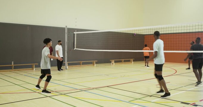 Diverse male players responding to initial serve, volleying pink purple volleyball over net in gym