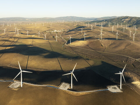 Aerial view of wind turbines standing tall against the rolling hills, a testament to renewable energy, bathed in the warm glow of the setting sun, Zahara de los Atunes, Andalusia, Spain.