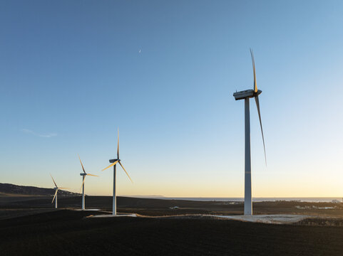 Aerial view of towering wind turbines piercing the horizon, their white forms stark against the deep blue sky and golden sunset glow, Zahara de los Atunes, Andalusia, Spain.