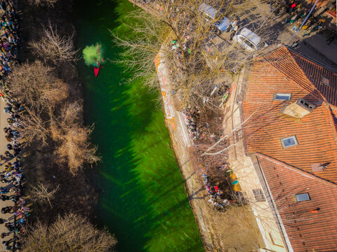 Aerial view of a vibrant green river flowing past aged brick buildings, with crowds gathering along the banks for St. Patrick's Day, Vilnius, Lithuania.