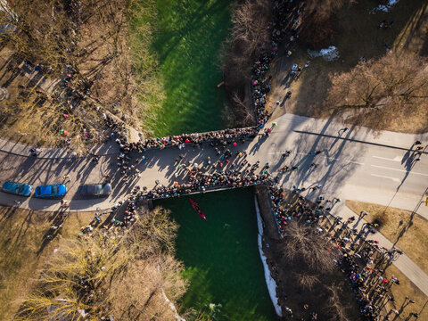 Aerial view of a bridge packed with revelers celebrating St. Patrick's Day, with the river dyed a vibrant green, a stark contrast to the surrounding bare trees and melting snow, Vilnius, Lithuania.