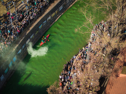 Aerial view of a vibrant green river reflecting the sky, with a small red boat cutting through the water as crowds gather along the stone banks, celebrating St. Patrick's Day, Vilnius, Lithuania.