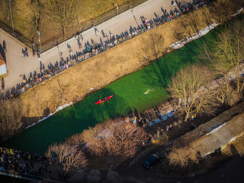 Aerial view of a vibrant green river flowing between bare trees and crowds of people, with a red boat cutting through the water, Vilnius, Lithuania.