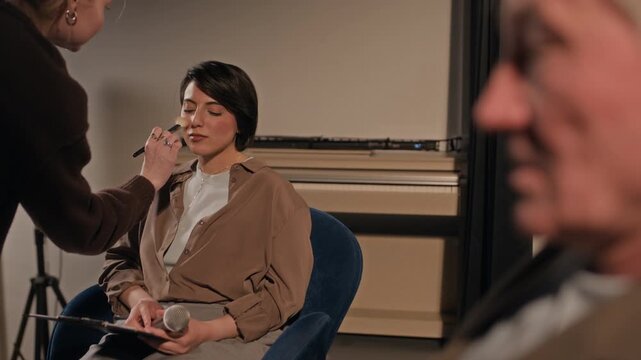 Medium shot of laughing multiethnic young female comedian chatting with colleagues backstage while reading through speech script on clipboard, artist applying makeup with face brush