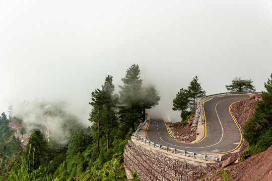 Aerial view of a winding road cutting through the lush green landscape, disappearing into the misty mountains, Pir Chanasi, Azad Kashmir, Pakistan.