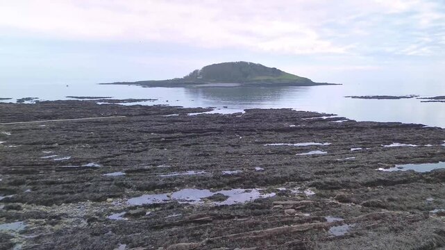 Aerial drone footage slowly flying towards Looe Island from Hannafore in Looe, Cornwall. Calm and peaceful sea with rocky shoreline in foreground transitioning into open water.