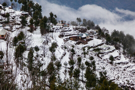 Aerial view of winter's touch paints the landscape in stark contrasts, with snow-laden roofs against the dark forest, Pir Chanasi, Azad Kashmir, Pakistan.