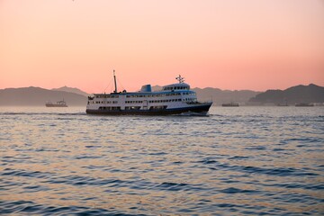 Obraz premium A classic white and blue Star Ferry sails across the water as the sky turns pink behind the distant mountain silhouettes