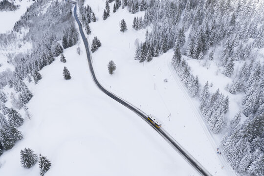 Aerial view of a winding road cuts through a snow-laden landscape, contrasting the dark asphalt with the pristine white snow and the dense green of the coniferous trees, Ormont-Dessus, Switzerland.